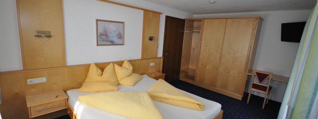 A cozy bedroom with a large double bed and yellow cushions. The walls are made of light wood and there is a television as well as a wardrobe. - Appartement Harmonie - Sölden