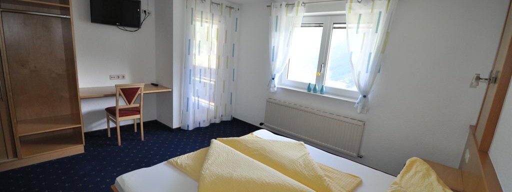 A simple and bright bedroom with a large bed, two pillows, and a wooden wardrobe. There is also a table with a chair and a window with curtains. - Appartement Harmonie - Sölden