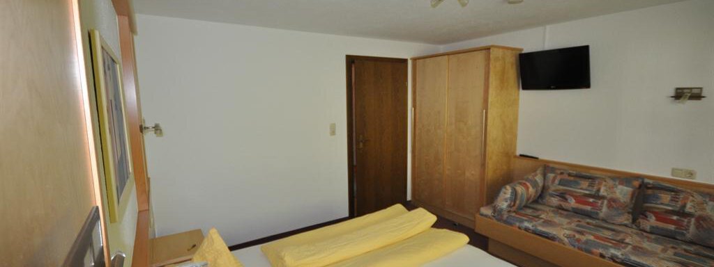 A simple bedroom with a double bed and a sofa. The walls are neutral, and a television is mounted on the wall. - Appartement Harmonie - Sölden