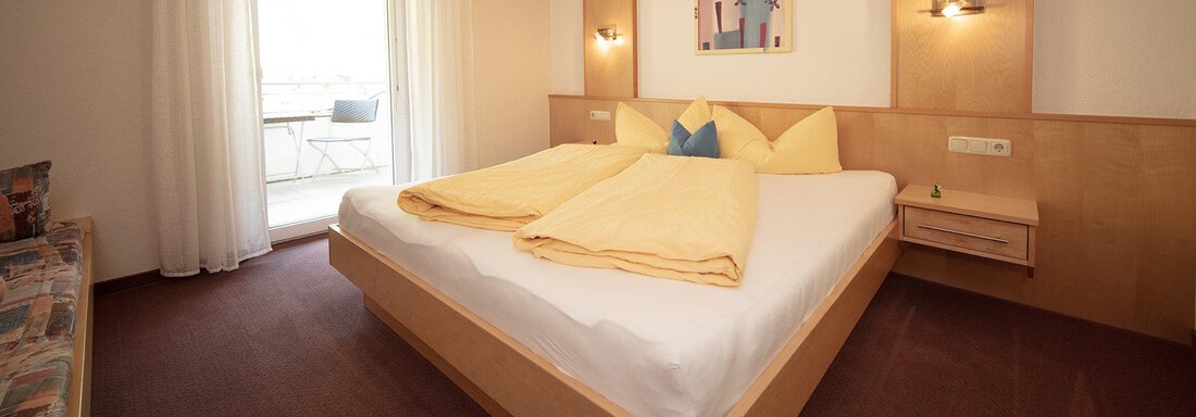 A modern bedroom with a large bed and yellow pillows. There are windows with curtains that let in daylight. - Appartement Harmonie - Sölden