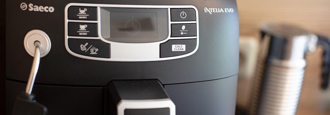 A modern coffee machine with a user-friendly display and various buttons. In the background, there is a milk frother. - Appartement Harmonie - Sölden
