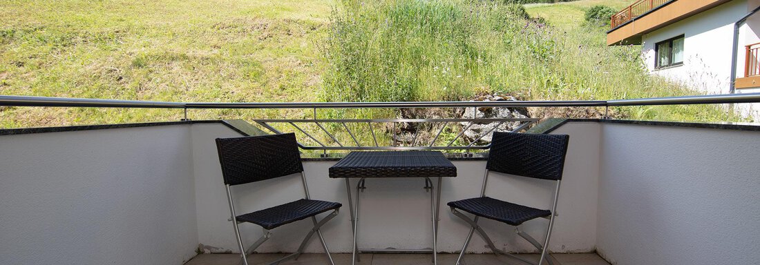A balcony with two black chairs and a table. In the background, green hills and a blue sky can be seen. - Appartement Harmonie - Sölden