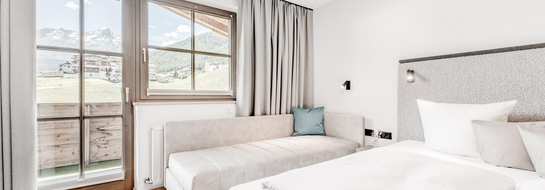 A modern hotel room with a large window and a view of the mountains. There is a double bed and a cozy seating area. - Appartement Montanara - Sölden