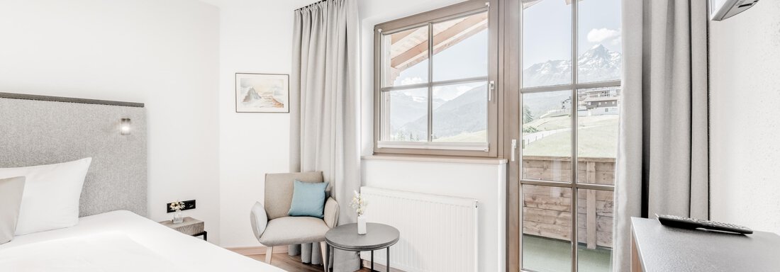 A modern bedroom with a large bed and bright curtains. The room has a small table and a beautiful view of the balcony. - Appartement Montanara - Sölden