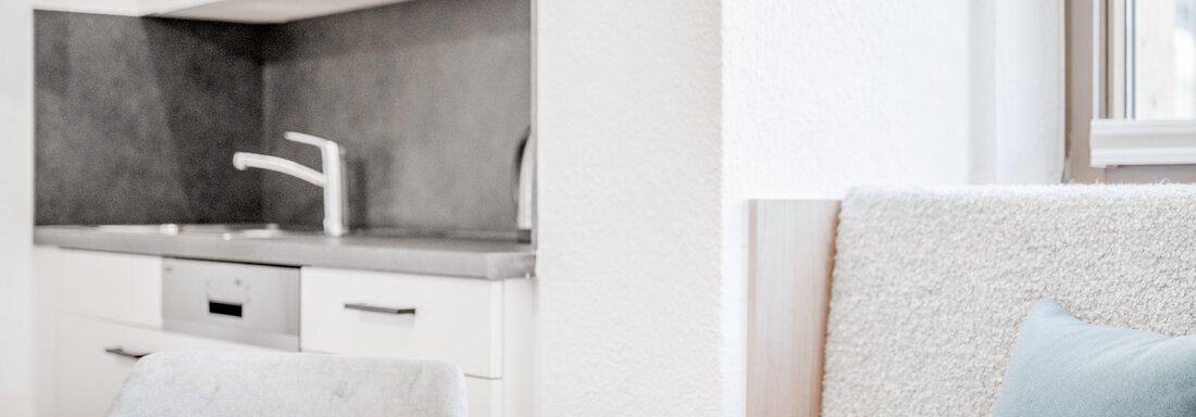 A modern, minimalist kitchen with light cabinets and a large window. The table is made of wood and next to it is a comfortable seating area. - Appartement Montanara - Sölden