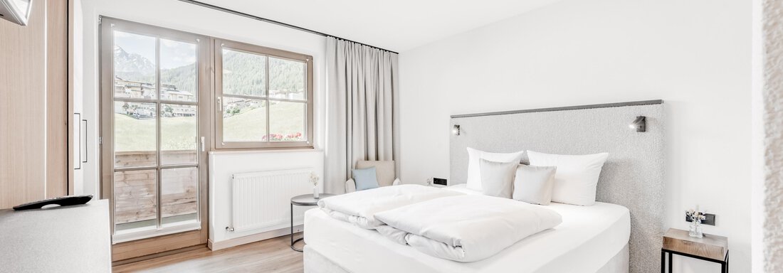 A modern bedroom with a large bed and bright walls. The room has windows with a view and minimalist decor. - Appartement Montanara - Sölden