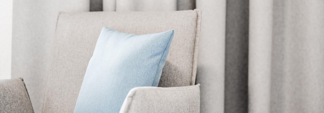 A cozy armchair with a blue cushion stands in front of a neutral curtain. The colors are soft and invite relaxation. - Appartement Montanara - Sölden