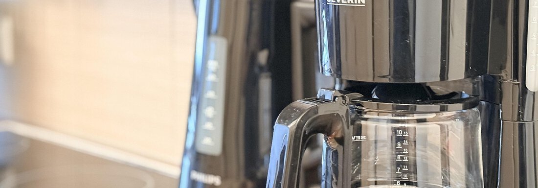 A coffee machine in black stands on a kitchen counter. In the background, a kettle can be seen. - Appartementhaus A CASA "Kristall" - Sölden