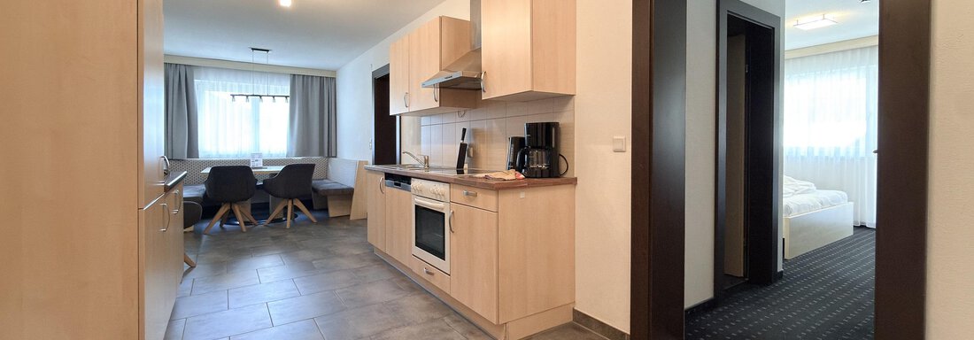 A modern kitchen with light wooden furniture and an open living area. In the background, a table with chairs and a bedroom are visible. - Appartementhaus A CASA "Kristall" - Sölden