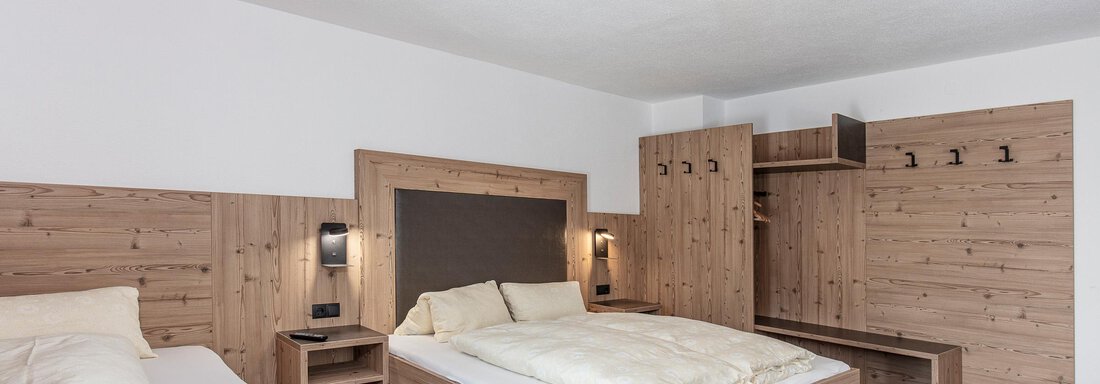 A cozy hotel room with two beds, a table, and a television. The walls are in warm wood tones and the carpet has a red pattern. - Appartementhaus Fender - Sölden