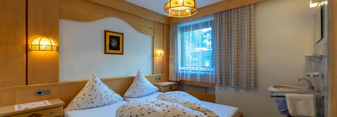 A cozy bedroom with a double bed and two pillows. The walls are made of light wood and there are windows with curtains as well as soft lighting. - Appartementhaus Karlinger - Sölden