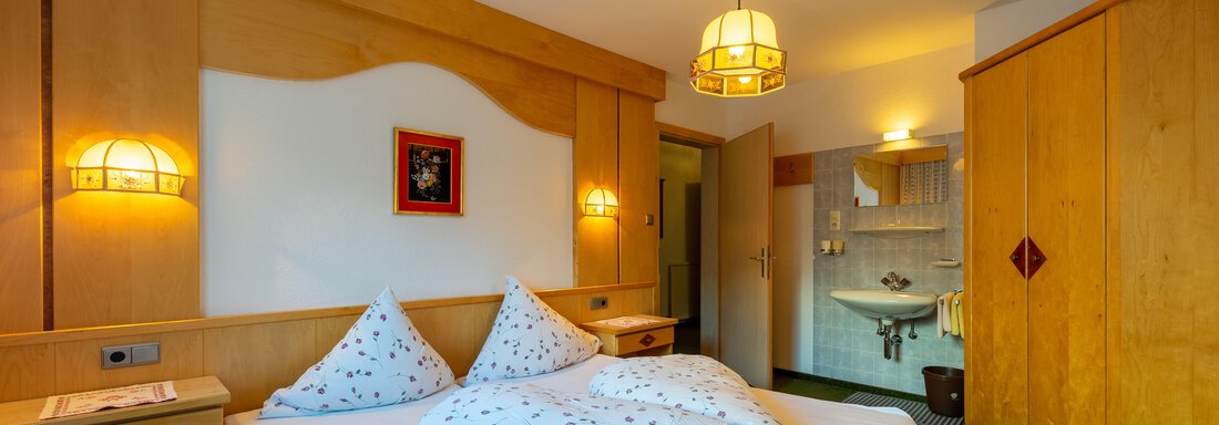 A cozy bedroom with a large bed and bright wooden furniture. In the background, a bathroom with modern sanitary facilities can be seen. - Appartementhaus Karlinger - Sölden