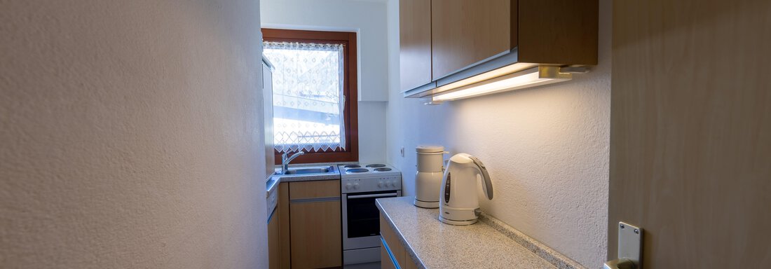A narrow, modern kitchen with wooden furniture and a window that lets in daylight. On the countertop, there is a kettle and a few kitchen appliances. - Appartementhaus Karlinger - Sölden