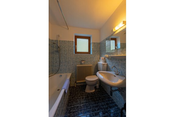 A modern bathroom with blue tiles and a shower over the bathtub. There is a sink, a toilet, and a window for natural light. - Appartementhaus Karlinger - Sölden