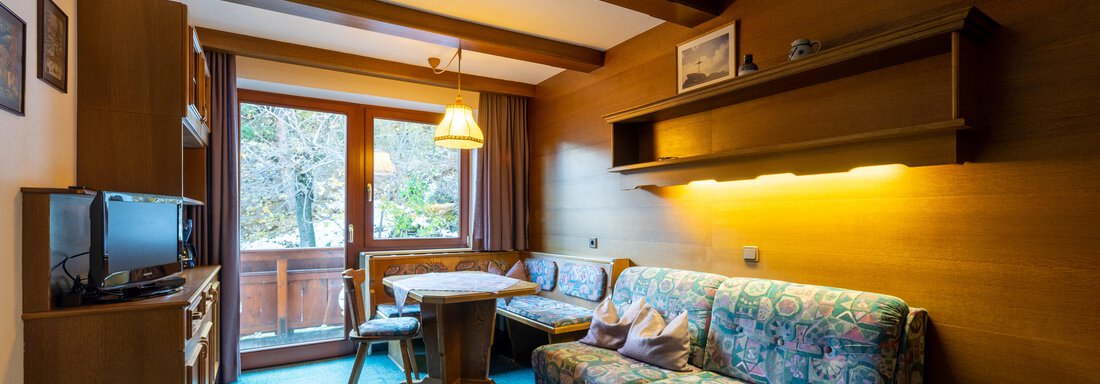 A cozy living room with a sofa, a table, and a television. Large windows offer a view of the nature outside. - Appartementhaus Karlinger - Sölden
