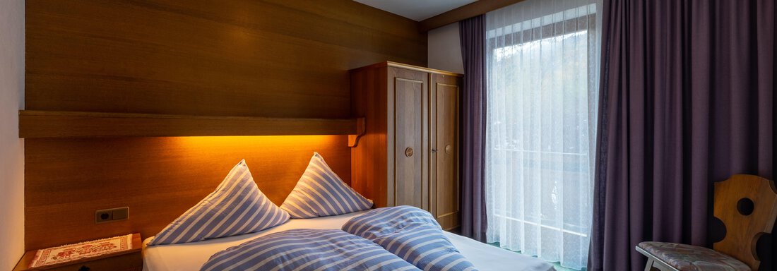 A cozy bedroom with a double bed and striped pillows. The room has windows with curtains and a wooden wardrobe. - Appartementhaus Karlinger - Sölden