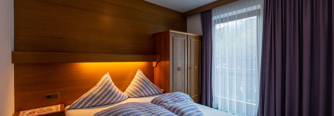 A cozy bedroom with a double bed and striped pillows. The room has a warm wood tone and large windows with curtains. - Appartementhaus Karlinger - Sölden