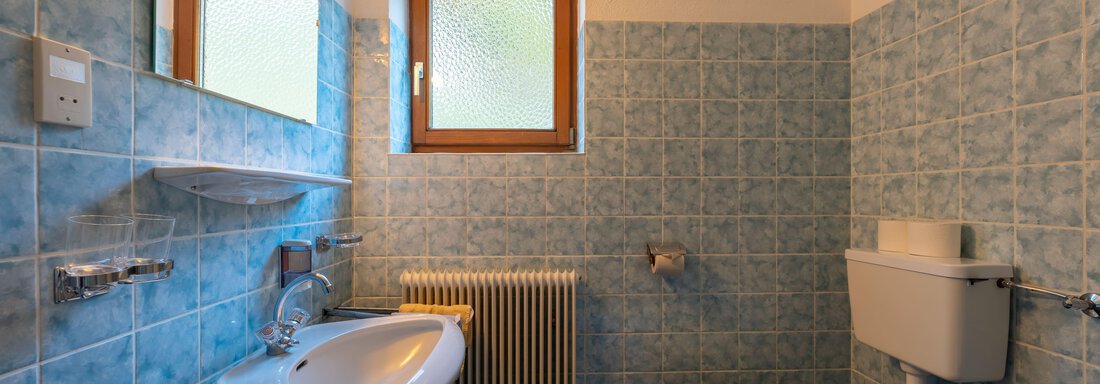 A small bathroom with blue tiles and a window. It features a sink, a toilet, and heating. - Appartementhaus Karlinger - Sölden