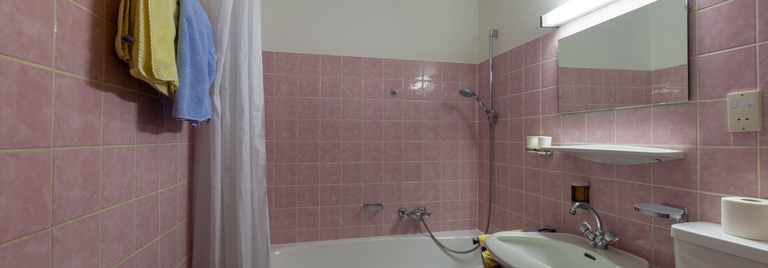 A small bathroom with pink tiles and a bathtub. Towels hang on the wall, and there is a sink and a mirror. - Appartementhaus Karlinger - Sölden
