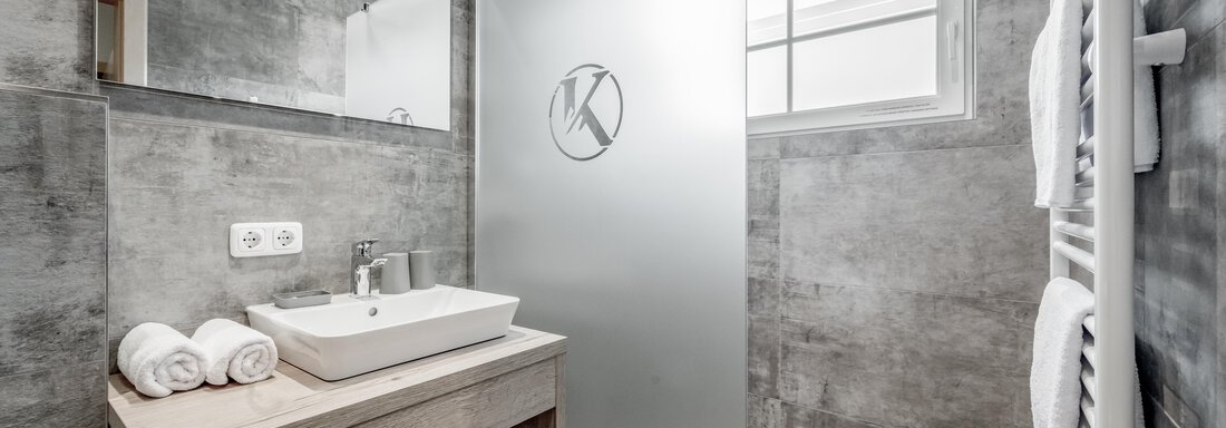 A modern bathroom with gray walls and a sink on a wooden cabinet. The shower is separated by a frosted glass panel. - Appartementhaus Kathrin - Sölden