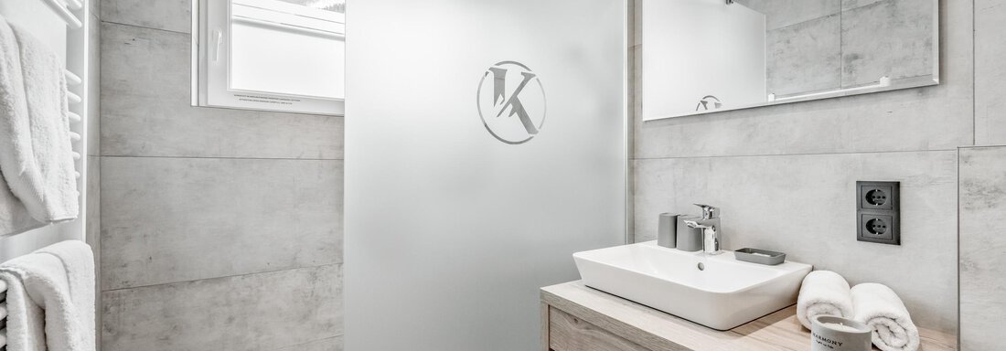 A modern bathroom with a shower-separating glass door. The space is in neutral shades of gray and features a wooden sink base. - Appartementhaus Kathrin - Sölden