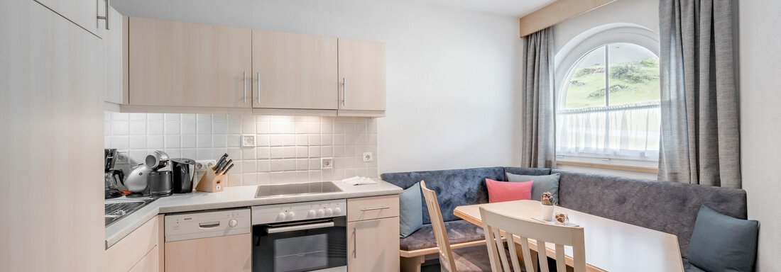 A modern kitchen with light wood cabinets and a dining area. The room has a large window and cozy seating. - Appartementhaus Kathrin - Sölden