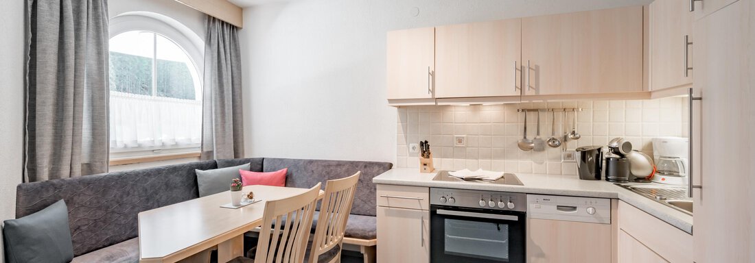 A modern kitchen with a dining area and a large window. The furniture is made of light wood and the walls are painted light. - Appartementhaus Kathrin - Sölden