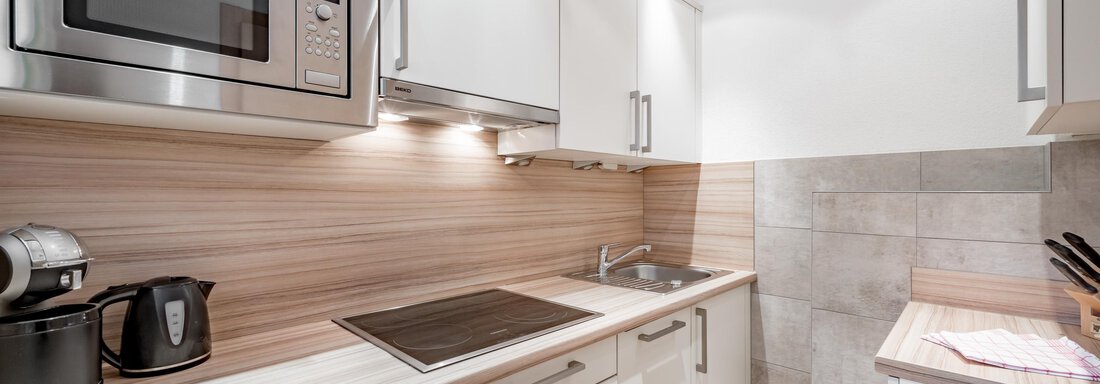 A modern kitchen with white cabinets and wood paneling. It is equipped with a microwave, a cooktop, and a sink. - Appartementhaus Kathrin - Sölden