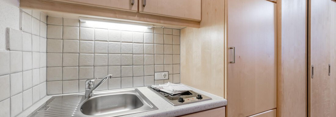 A modern kitchen unit made of wood with a sink and a small cooking area. The walls are tiled with white tiles, and there is plenty of storage space. - Appartementhaus Kathrin - Sölden