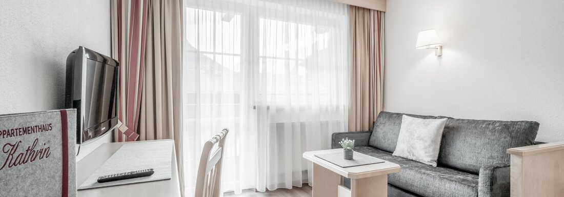A modern living room with a gray sofa and a table. The large windows let in a lot of light, and the room feels bright and inviting. - Appartementhaus Kathrin - Sölden