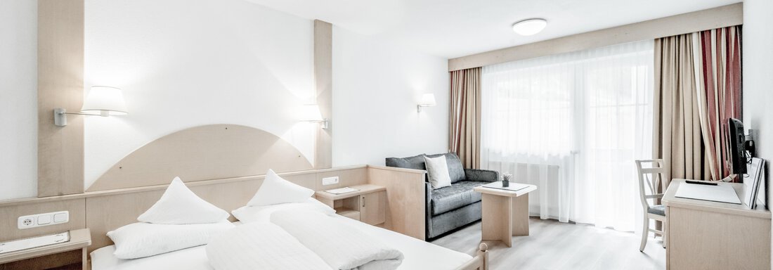 A modern room with a large bed, a sofa, and a desk. The space is bright and friendly with a window that lets in plenty of daylight. - Appartementhaus Kathrin - Sölden