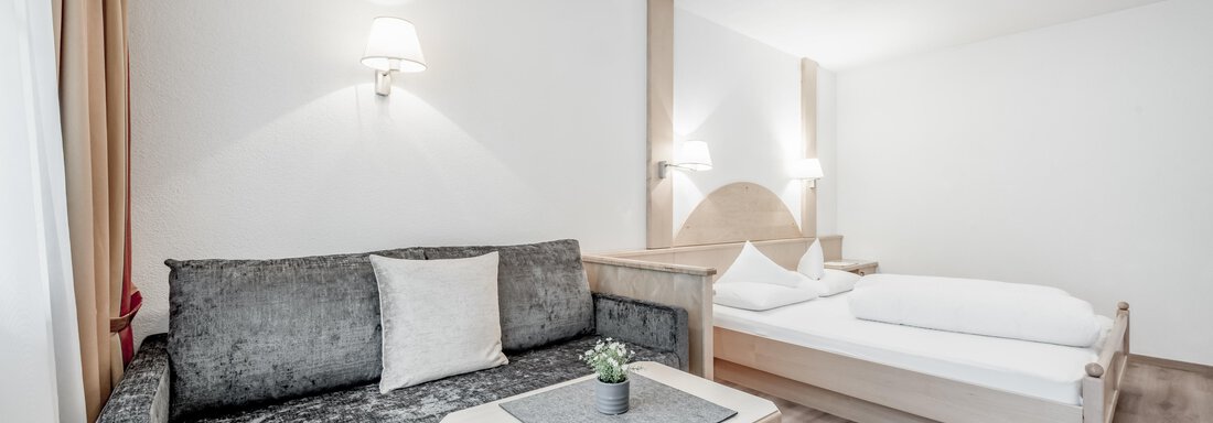 A bright and modern room with a comfortable sofa and a large bed. The walls are plain and the lighting creates a cozy atmosphere. - Appartementhaus Kathrin - Sölden