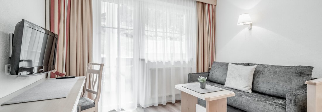 A cozy living room with a gray sofa and a small table. Bright curtains let in a lot of light. - Appartementhaus Kathrin - Sölden