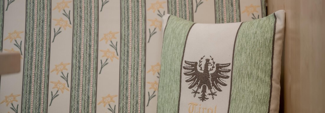 A cozy sofa with a cushion featuring the coat of arms of Tyrol. The colors are green and beige, with a floral pattern in the background. - Appartementhaus Kathrin - Sölden