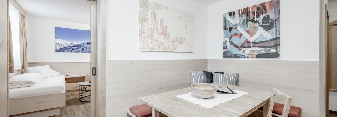 A cozy dining area with a wooden table and chairs. In the background, a bedroom with a bed and a window is visible. - Appartementhaus Kathrin - Sölden