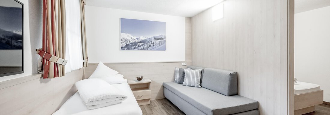 A modern room with bright walls and a wooden floor. It contains a single bed, a couch, and a picture of a mountain landscape on the wall. - Appartementhaus Kathrin - Sölden