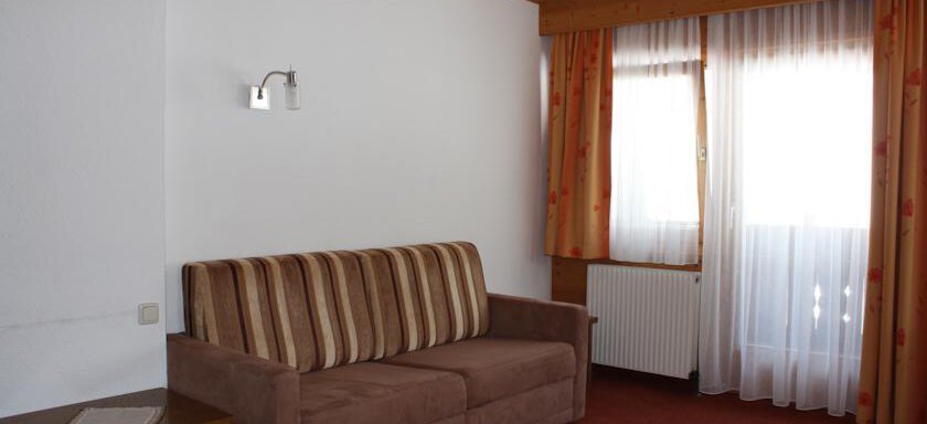 A cozy living room with a beige sofa and a table. The windows are decorated with white curtains and orange drapes, and the room features warm wooden construction. - Appt. Magda - Sölden