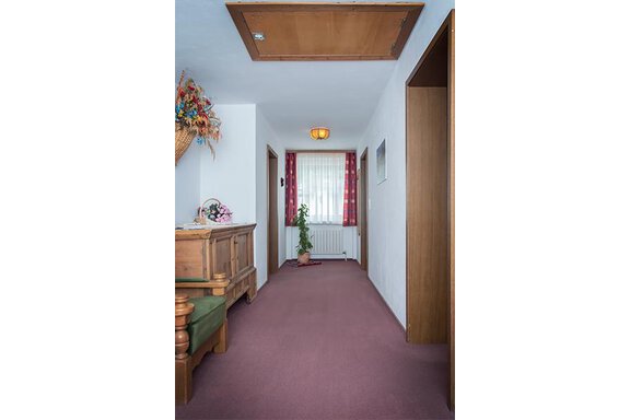 A bright hallway with a carpet in purple and wooden furniture. At the end of the hallway, there is a plant in front of a window with curtains. - Appt. Schnöller - Sölden