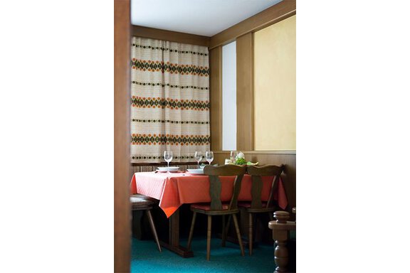 A cozy dining area with an orange table and wooden chairs. The walls are decorated with a decorative curtain and a warm hue. - Appt. Schnöller - Sölden