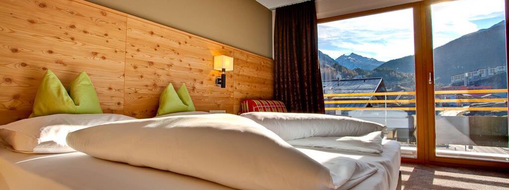 Ein gemütliches Zimmer mit einem großen Bett und zwei grünen Kissen. Durch das Fenster ist ein schöner Blick auf die Berge zu sehen. - Bäckelar Wirt - Sölden