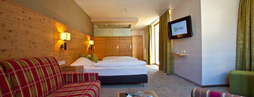 Ein modernes Hotelzimmer mit Holzwänden und einem gemütlichen Schlafbereich. Es gibt ein Sitzbereich mit kariertem Stoff und große Fenster, die viel Licht hereinlassen. - Bäckelar Wirt - Sölden