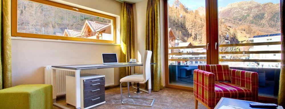 Ein modernes Zimmer mit einem Schreibtisch, einem bequemen Stuhl und einem bunten Sessel. Durch die großen Fenster hat man einen Blick auf die Berge und den Außenbereich. - Bäckelar Wirt - Sölden