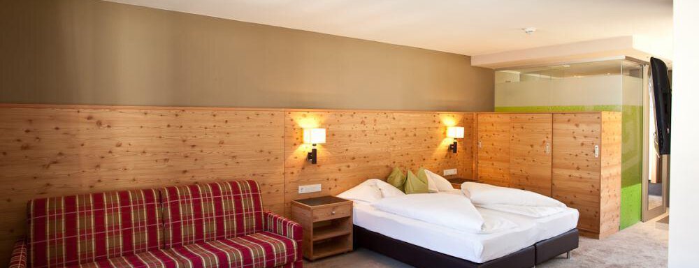 Ein gemütliches Hotelzimmer mit einem Doppelbett und einem roten karierten Sofa. Die Wände sind teilweise aus Holz, was eine warme Atmosphäre schafft. - Bäckelar Wirt - Sölden