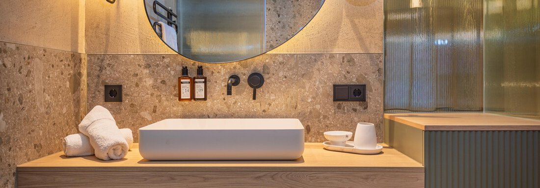 Ein modernes Badezimmer mit einem eleganten Waschtisch und einem großen Spiegel. Die Wände haben eine stilvolle Steinoptik, und es gibt frische Handtücher sowie Pflegeprodukte. - Bäckelar Wirt - Sölden