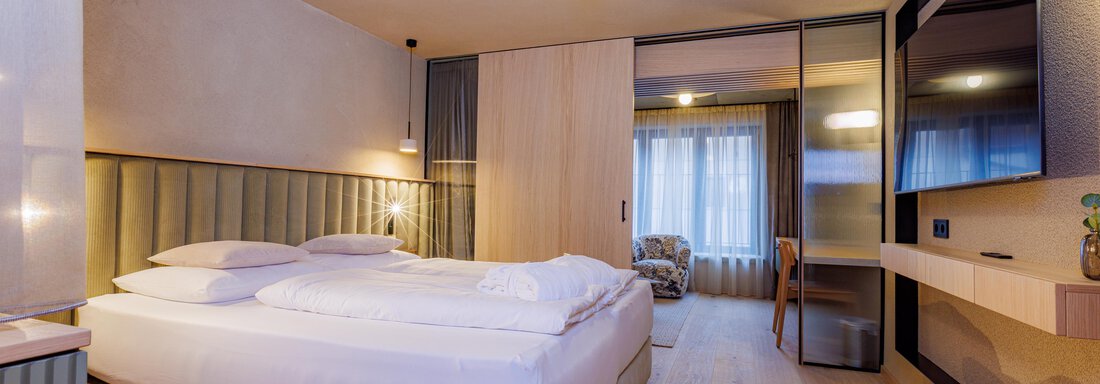 Ein modernes Hotelzimmer mit einem großen Bett und einem klaren, minimalistischen Design. Der Raum hat Holzfußböden und viel natürliches Licht von einem Fenster. - Bäckelar Wirt - Sölden