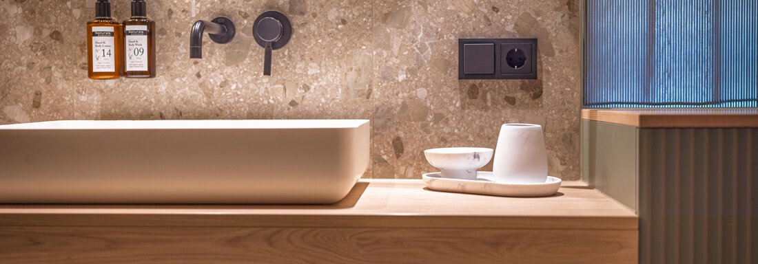 Ein modernes Badezimmer mit einer stilvollen Holzwaschtischplatte und einem eleganten Waschbecken. Die beige Steinwand und die runden Spiegel verleihen dem Raum ein gemütliches Ambiente. - Bäckelar Wirt - Sölden