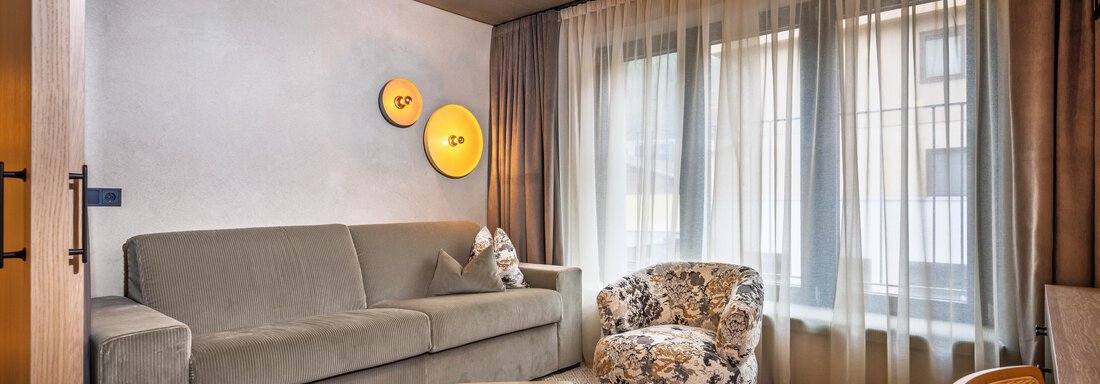 Ein modernes Wohnzimmer mit einem grauen Sofa und einem gepolsterten Sessel. Große Fenster lassen viel Tageslicht herein und verleihen dem Raum eine einladende Atmosphäre. - Bäckelar Wirt - Sölden