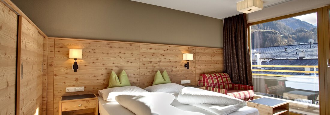 Ein modernes Hotelzimmer mit einem Doppelbett, einer gemütlichen Sitzecke und Holzverkleidungen. Das große Fenster bietet einen Blick auf die Berge. - Bäckelar Wirt - Sölden