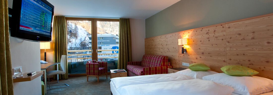 Ein modernes Hotelzimmer mit zwei Betten, einem Fernseher und einem gemütlichen Sitzbereich. Durch die Fenster ist eine wunderbare Aussicht auf die Berge sichtbar. - Bäckelar Wirt - Sölden