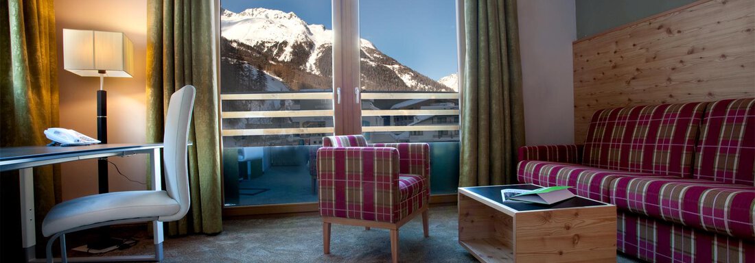 Ein gemütliches Hotelzimmer mit Blick auf schneebedeckte Berge. Die Einrichtung besteht aus einem Sofa, einem Tisch und einem Schreibtisch. - Bäckelar Wirt - Sölden